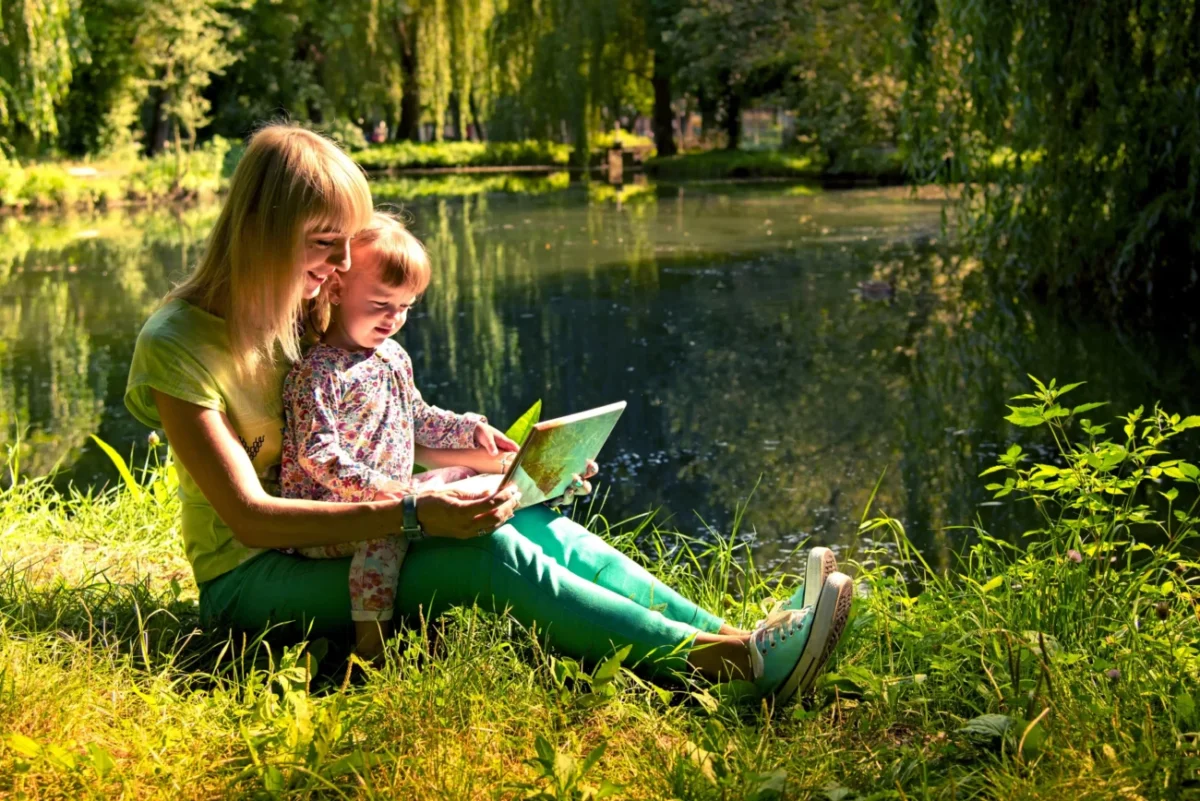 Žena s holčičkou sedí v trávě u vody a čtou pohádky.