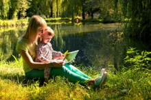 Žena s holčičkou sedí v trávě u vody a čtou pohádky.