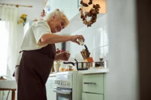 úhledná babička v hnědé zástěře (bílá košile, šedé vlasy) u linky v moderní kuchyni dává koření do polévky v nerez hrnci