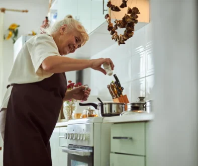 úhledná babička v hnědé zástěře (bílá košile, šedé vlasy) u linky v moderní kuchyni dává koření do polévky v nerez hrnci