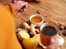 detail na přípravu zázvorového čaje - ženská ruka ve žlutém svetru drží lžičku s medem, na dřevěném stole hrnek, citron, zázvor na tácku