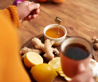 detail na přípravu zázvorového čaje - ženská ruka ve žlutém svetru drží lžičku s medem, na dřevěném stole hrnek, citron, zázvor na tácku
