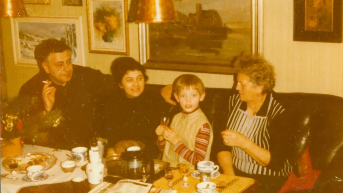 Retro foto ze 70.-80. let. Rodinná party v obýváku (5 lidí na pohovce u stolku, za nimi na zdech obrazy)