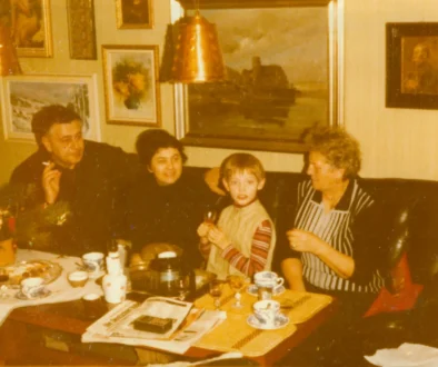 Retro foto ze 70.-80. let. Rodinná party v obýváku (5 lidí na pohovce u stolku, za nimi na zdech obrazy)