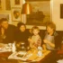 Retro foto ze 70.-80. let. Rodinná party v obýváku (5 lidí na pohovce u stolku, za nimi na zdech obrazy)