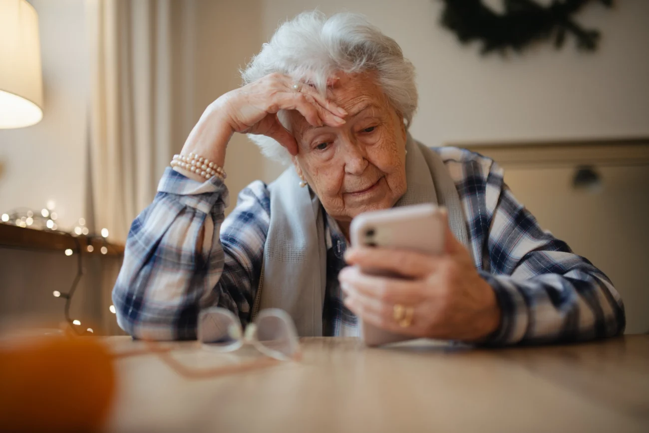 Stará žena sedí u stolu a kouká do mobilního telefonu. 