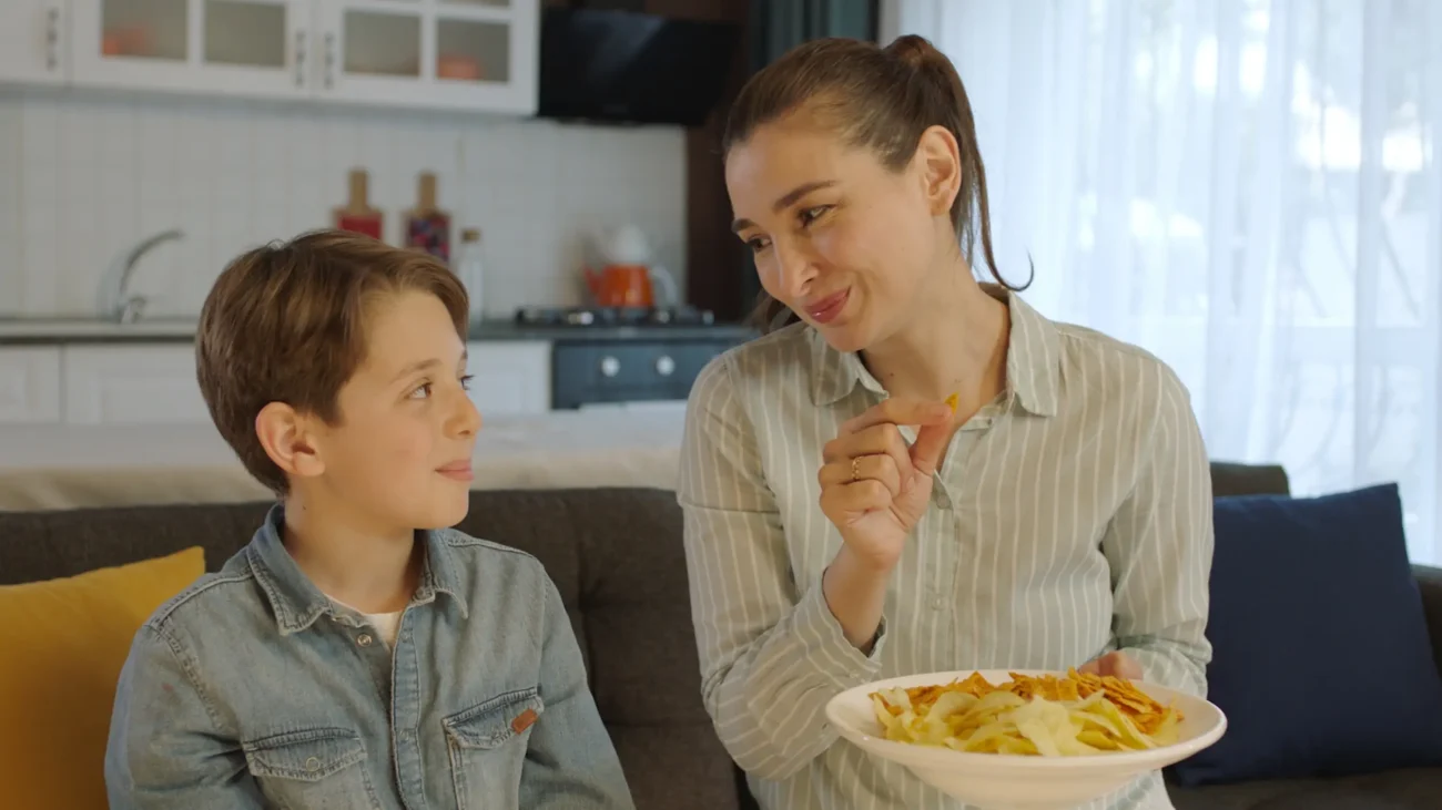 shutterstock_2252591457-2 | TVGURU.cz Mladá žena v ruce drží chipsy a sedí vedle chlapce na gauči.