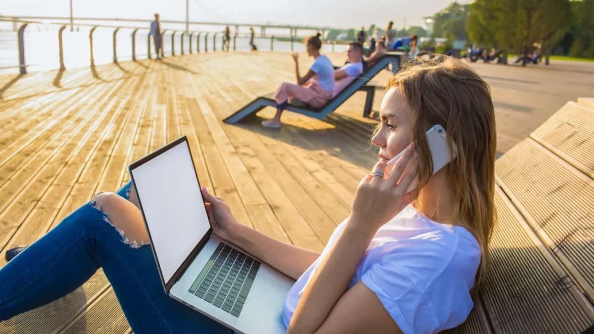 shutterstock_1235191966-2 Žena leží u pláže s notebookem na nohách a mobilem v ruce.