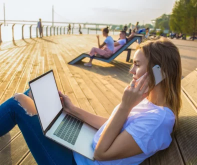 shutterstock_1235191966-2 Žena leží u pláže s notebookem na nohách a mobilem v ruce.