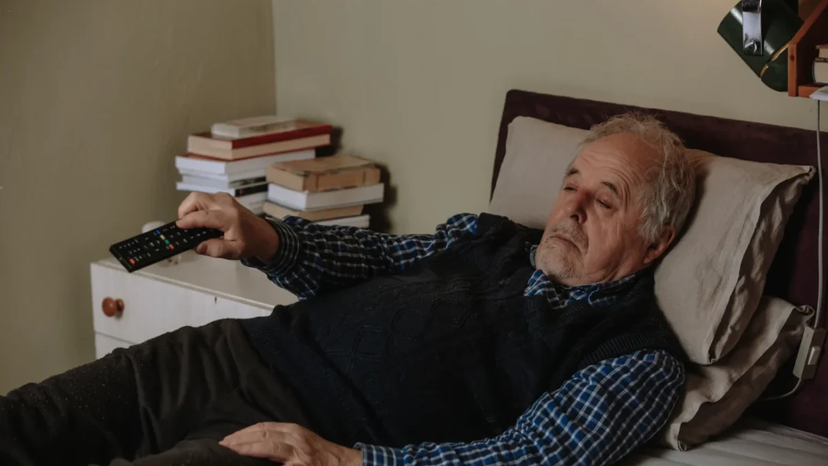 Muž leží na posteli s ovladačem v ruce.