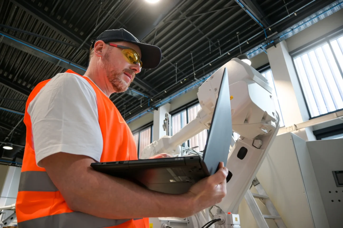 Muž stojí s notebookem v ruce u stroje. 