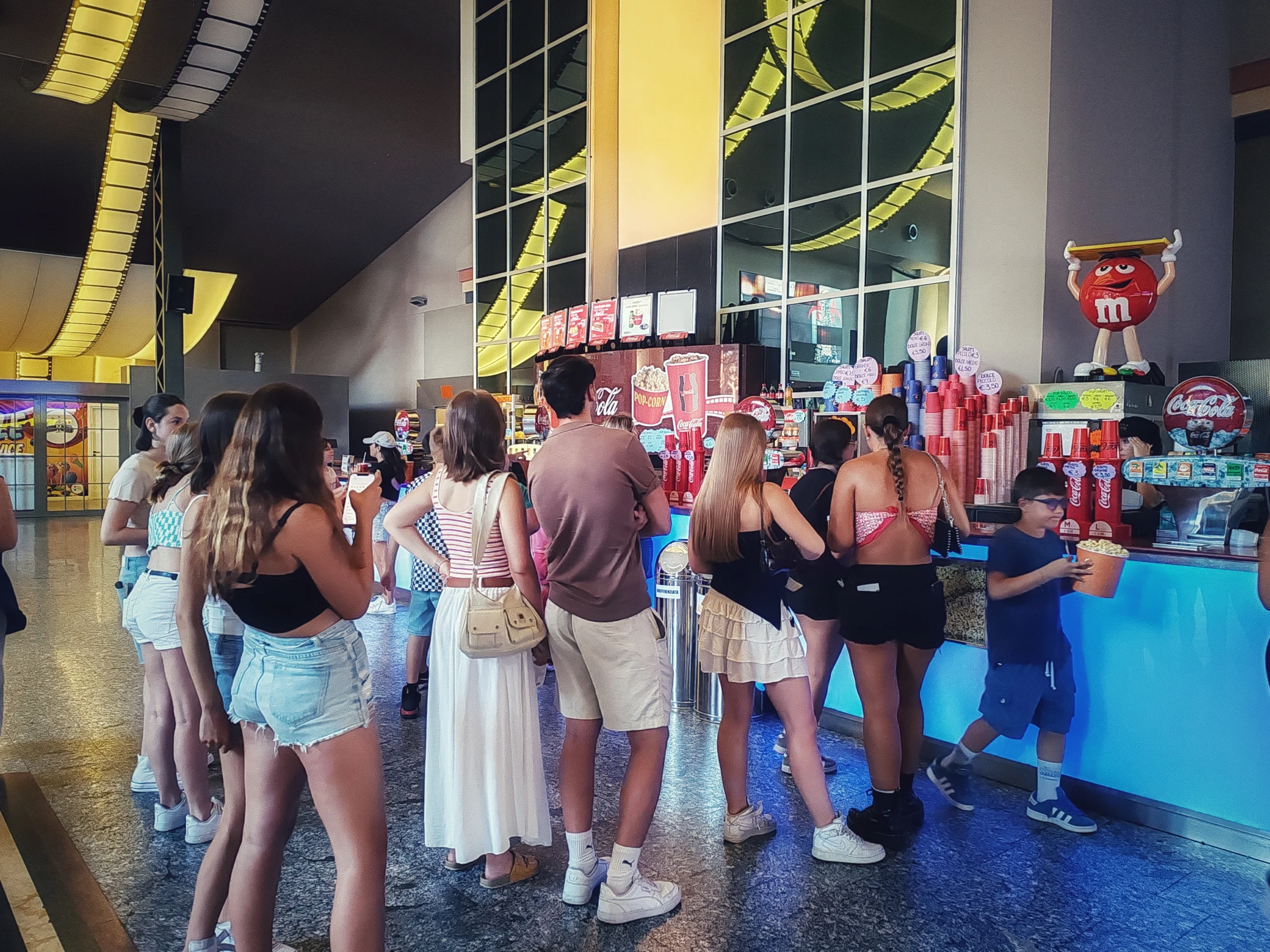 Řím, 30. června 2025. Řada lidí, hlavně dívek a chlapců, ve frontě na popcorn a další občerstvení v předsálí kina před promítáním filmu