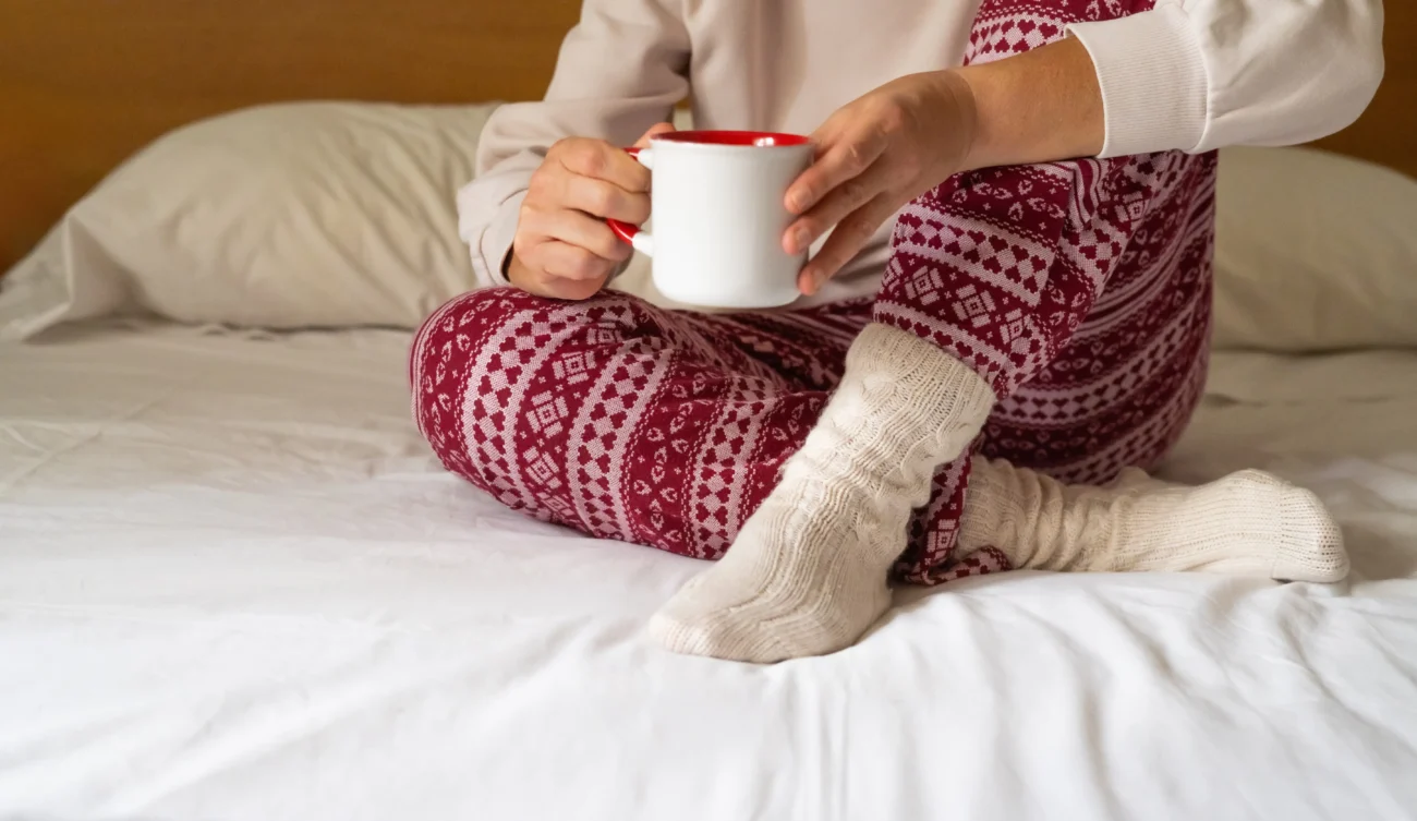 shutterstock_2528784359 1 | TVGURU.cz Žena sedící na posteli ve vánočním pyžamu s hrnkem v ruce.