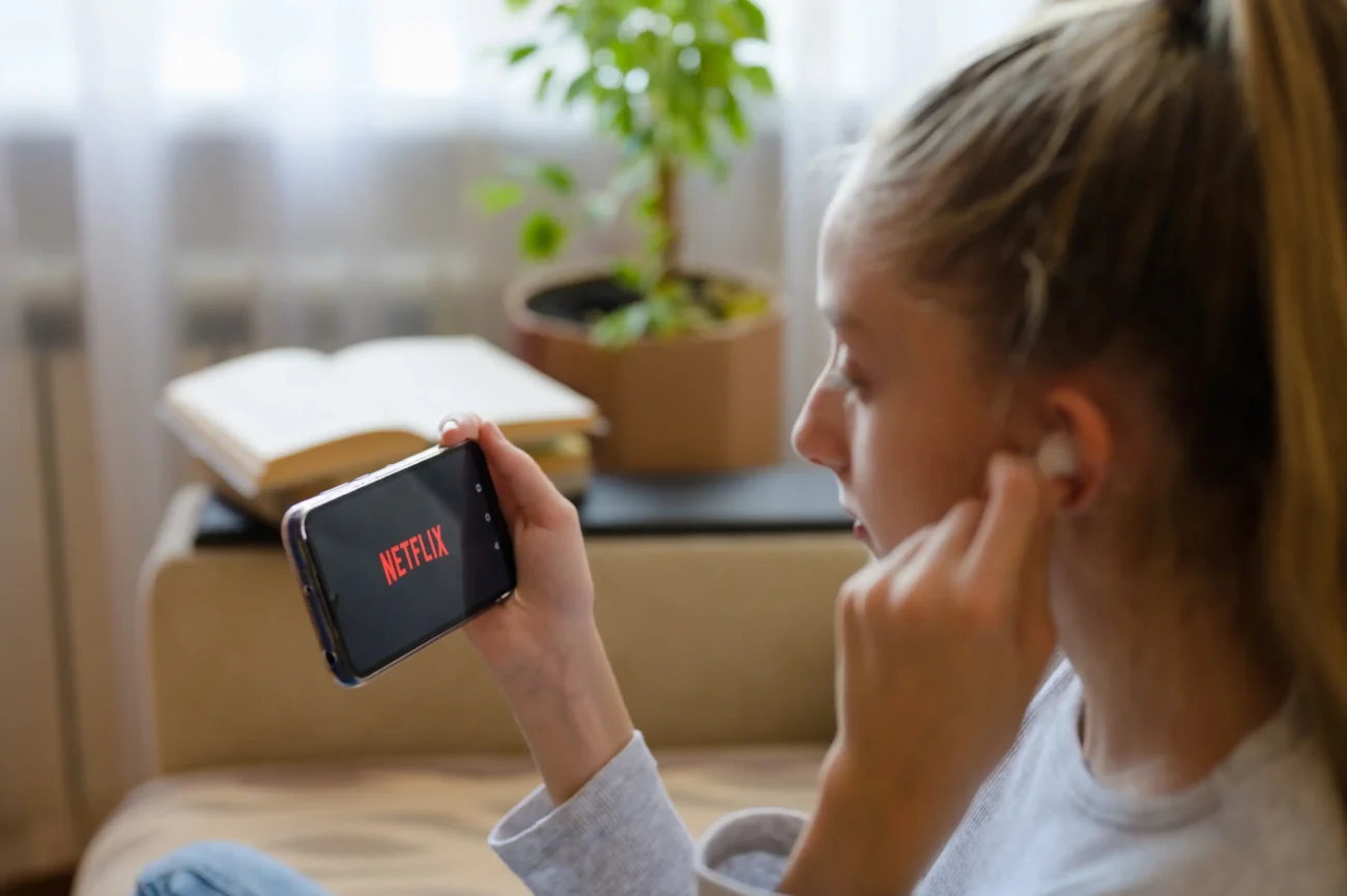 Slečna má na mobilu spuštěný Netflix a do uší si dává sluchátka.