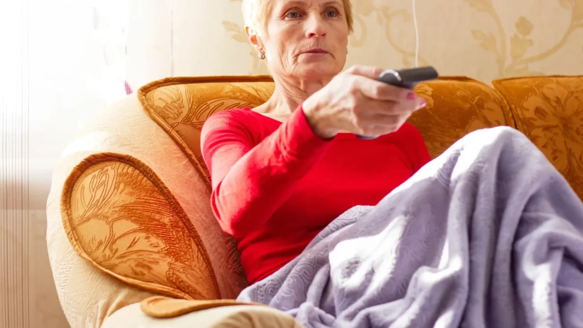 Babička sedí na gauči a míří na TV ovladačem.