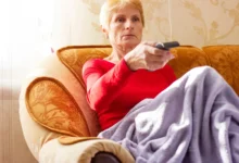 Babička sedí na gauči a míří na TV ovladačem.