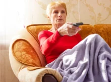 Babička sedí na gauči a míří na TV ovladačem.