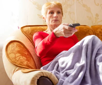 Babička sedí na gauči a míří na TV ovladačem.