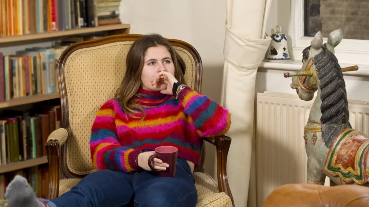 Slečna sedí ve svetru na křesle a kolem ní jsou knížky.