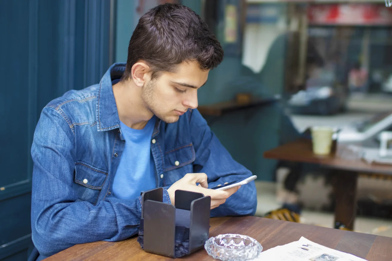 Mladík sedí u stolu a kouká na něco na telefonu. 