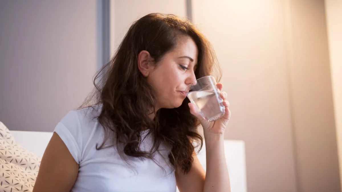 Žena sedí na posteli a popíjí vodu.