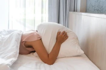 Žena leží v posteli a na hlavě má polštář.