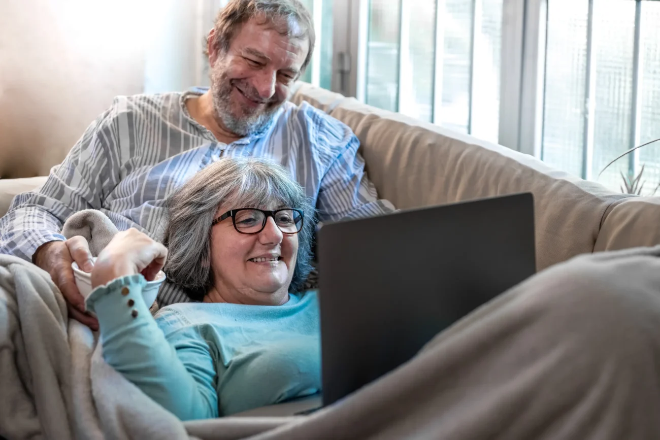 Děda s babičkou leží na gauči a dívají se na notebook. 