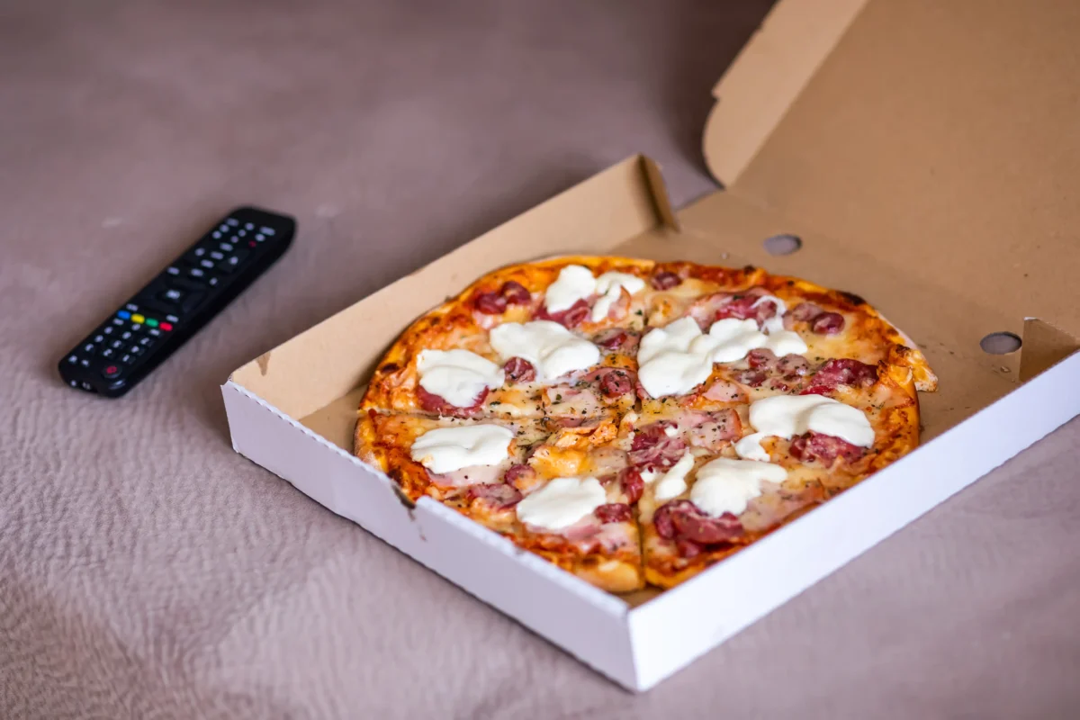 Na posteli leží krabice s pizzou a ovladač od TV.