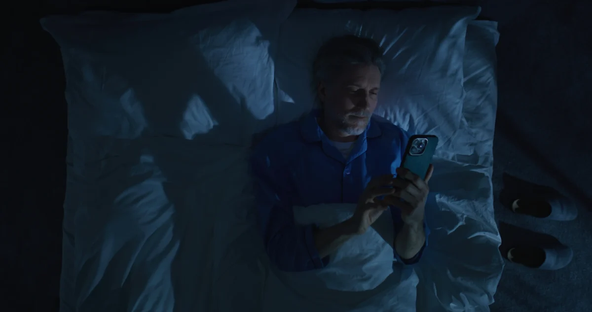 Člověk leží s mobilem v posteli. 