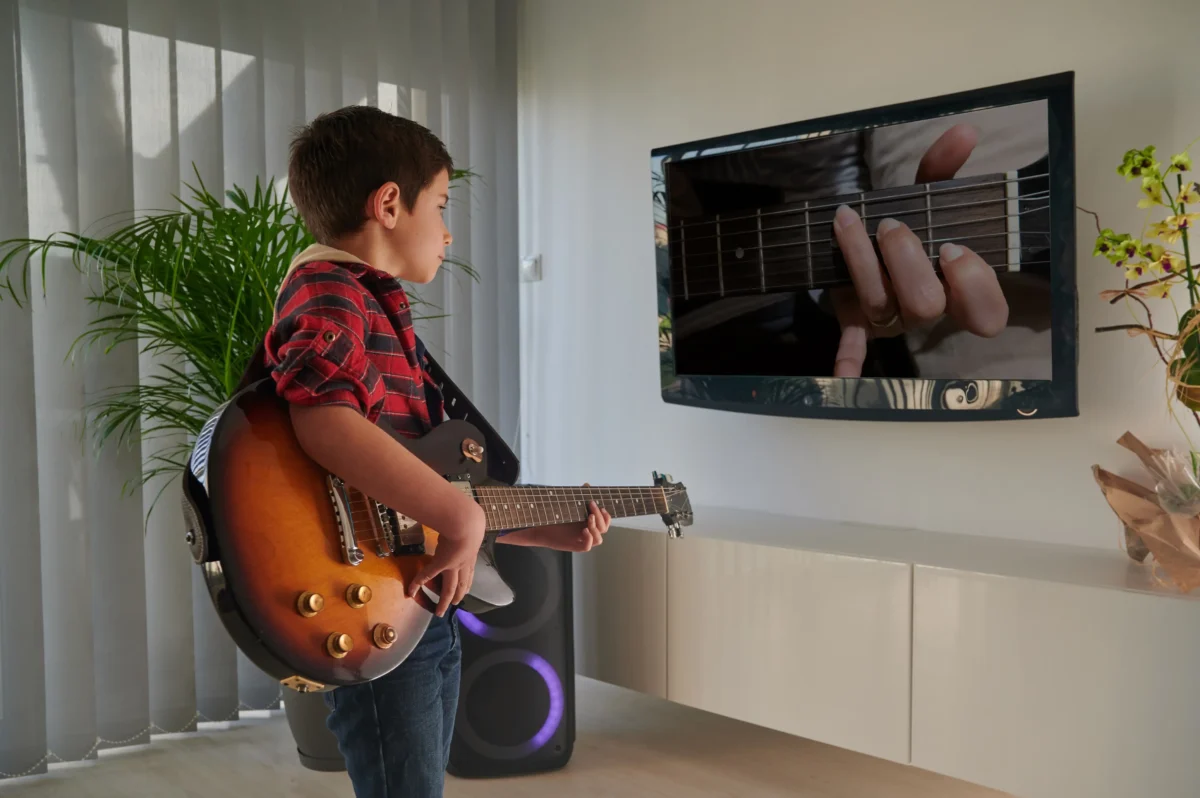 Chlapeček stojí před TV a hraje na kytaru. 