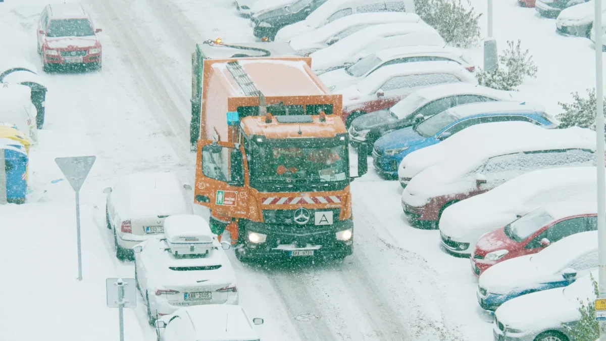 Popeláři projíždějí mezi zasněženými auty.