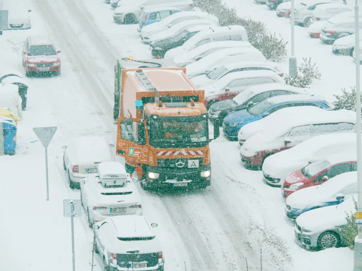 Popeláři projíždějí mezi zasněženými auty.