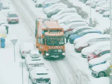 Popeláři projíždějí mezi zasněženými auty.