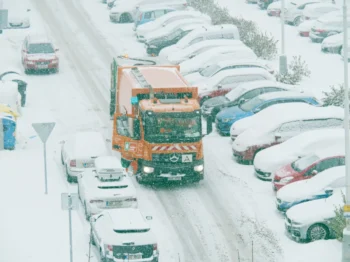 Popeláři projíždějí mezi zasněženými auty.
