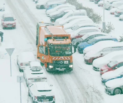 Popeláři projíždějí mezi zasněženými auty.
