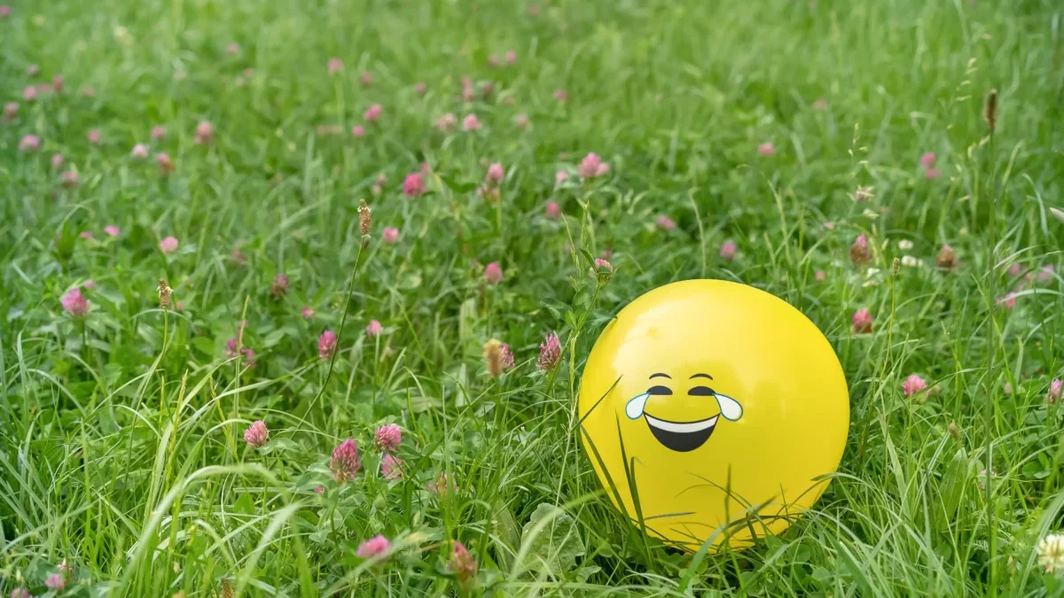 V trávě se válí balonek s nakresleným úsměvem.
