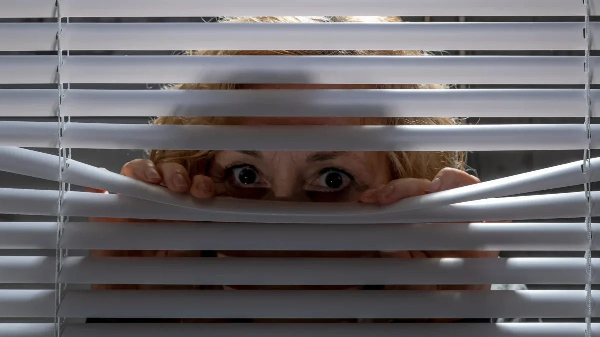 Žena vykukuje za žaluziemi.