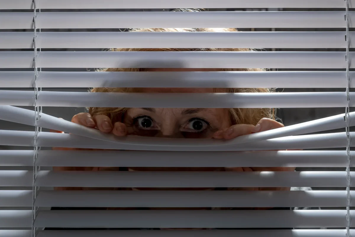 Žena vykukuje za žaluziemi.