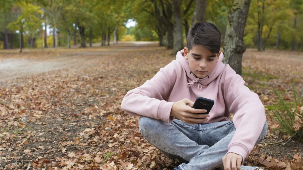 Chlapec sedí v přírodě na zemi a v ruce má telefon.
