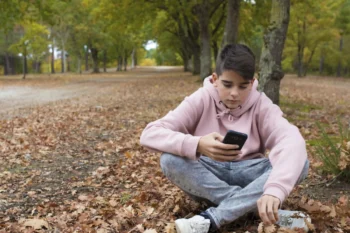 Chlapec sedí v přírodě na zemi a v ruce má telefon.