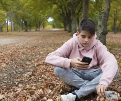 Chlapec sedí v přírodě na zemi a v ruce má telefon.