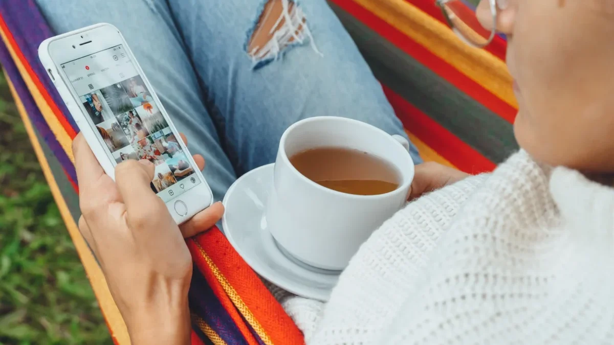 Žena projíždí na mobilu Instagram a má na klíně hrnek.