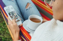 Žena projíždí na mobilu Instagram a má na klíně hrnek.