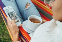 Žena projíždí na mobilu Instagram a má na klíně hrnek.