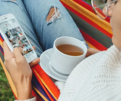 Žena projíždí na mobilu Instagram a má na klíně hrnek.