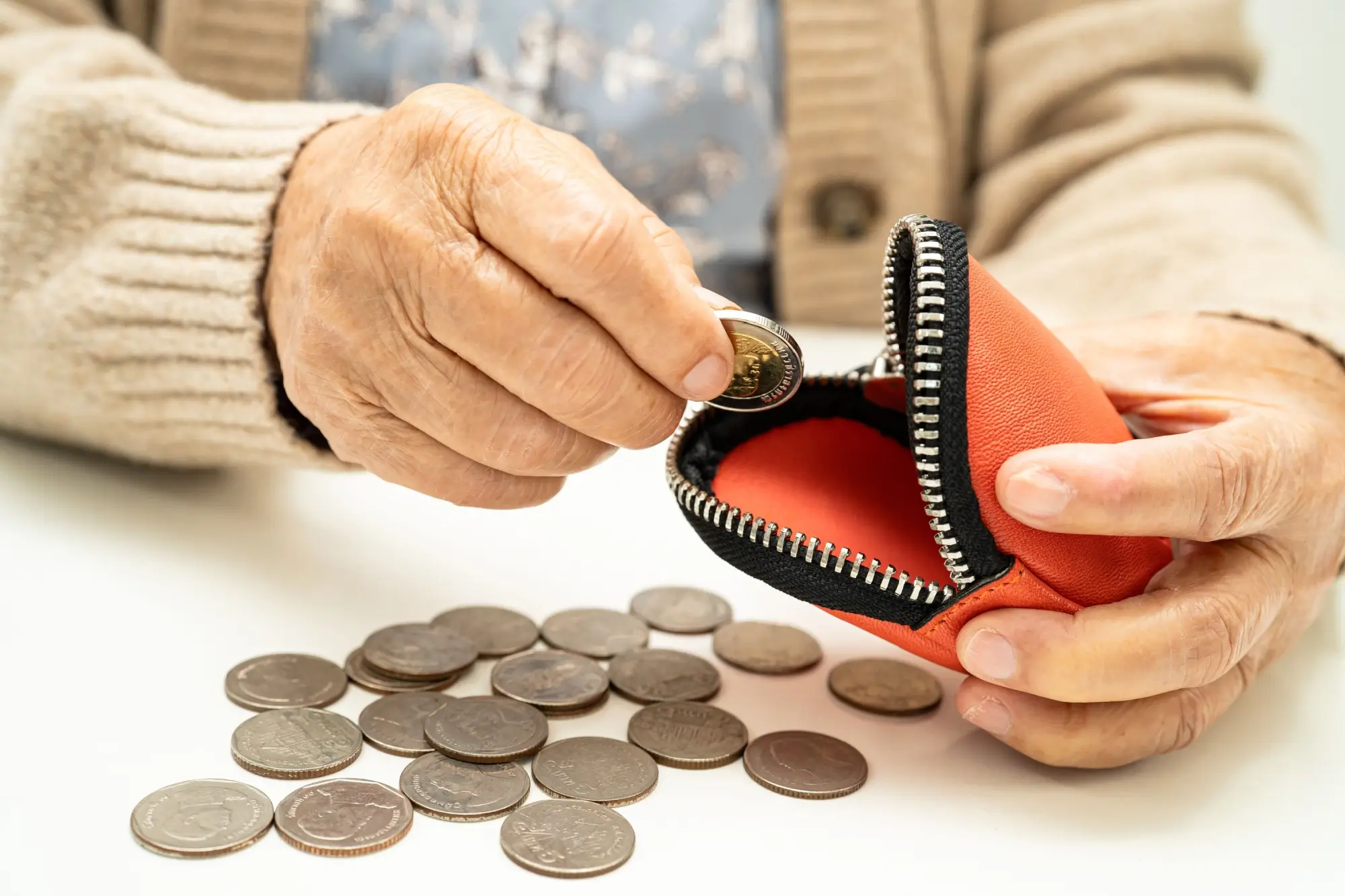 Close-up of an elderly person counting coins with an orange zippered wallet on a white surface nearby.