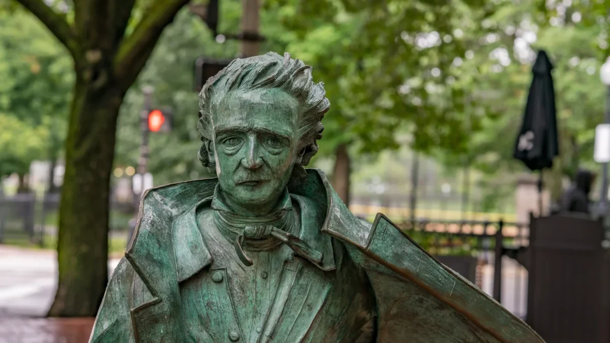 Bronze-green statue of a man in a suit with wild hair, holding a violin against a park backdrop of trees and benches.