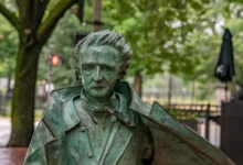 Bronze-green statue of a man in a suit with wild hair, holding a violin against a park backdrop of trees and benches.