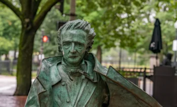 Bronze-green statue of a man in a suit with wild hair, holding a violin against a park backdrop of trees and benches.