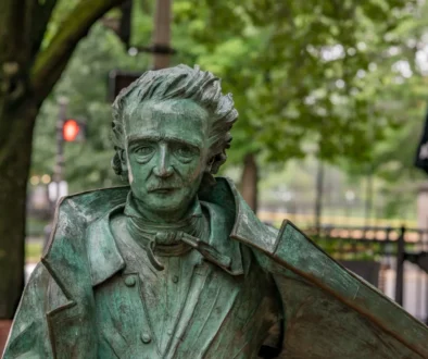 Bronze-green statue of a man in a suit with wild hair, holding a violin against a park backdrop of trees and benches.